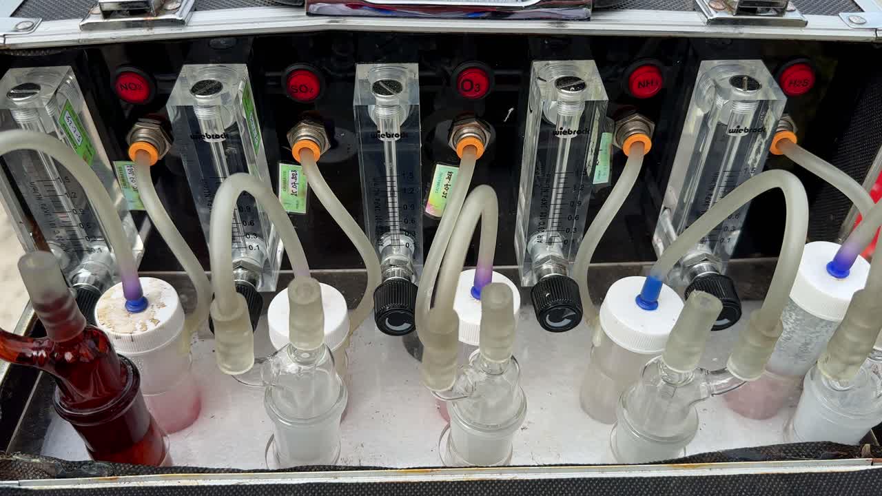 Close-up video of a precision fluid measurement system in a laboratory setup, showing indicators, connectors, and transparent hoses. Impinger machine for checking air quality.