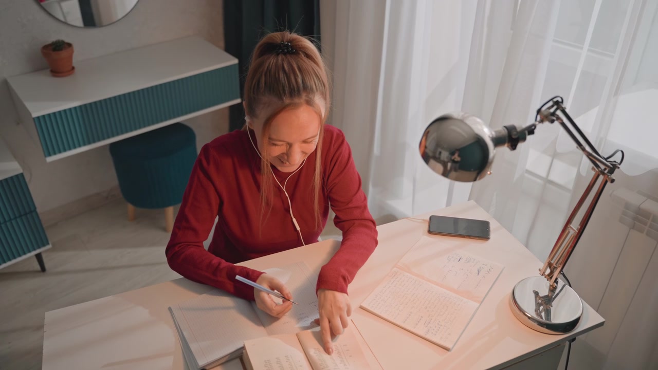 Mladá žena v červeném tričku s dlouhým rukávem studuje u stolu a píše. Na stole je linkovaný papír a chytrý telefon na kovové nabíječce. Scéna zůstává konzistentní, zatímco se žena soustředí na studijní materiály.