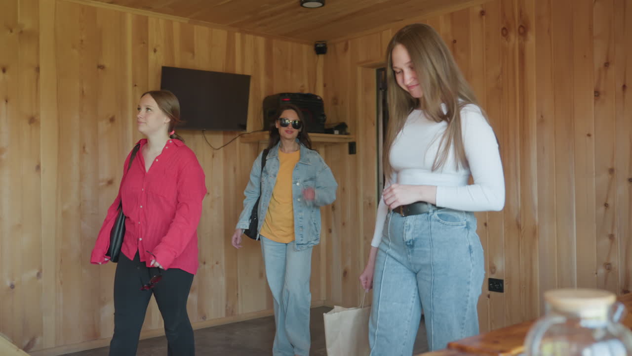 Glass jar and candle holders rest on wooden table in cozy cabin setting as three sisters walk into room in background smiling and looking around with curiosity and excitement