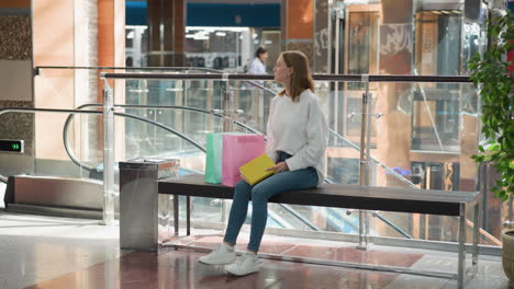 young woman seated on bench in modern shopping mall holding yellow book looks away thoughtfully, surroundings include colorful shopping bags, glass railings, escalator, and potted plants