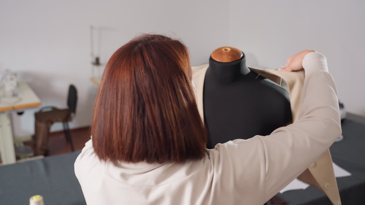 Fashion designer puts beige sleeveless blazer onto black dress form, carefully adjusting blazer into correct position, fastening front button, aligning seams and shoulders with precision