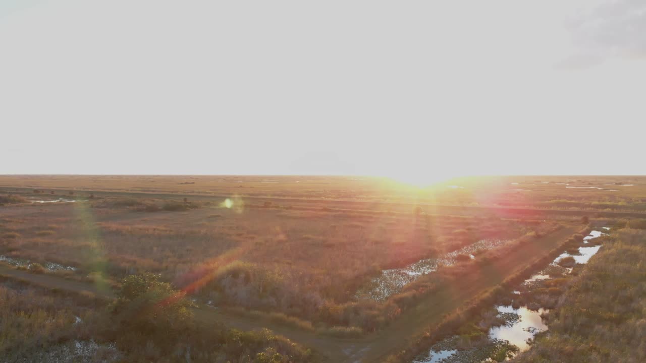 Aerial dolly-forward shot moving into bright sunlight over a wide wetland nature preserve with scattered water channels