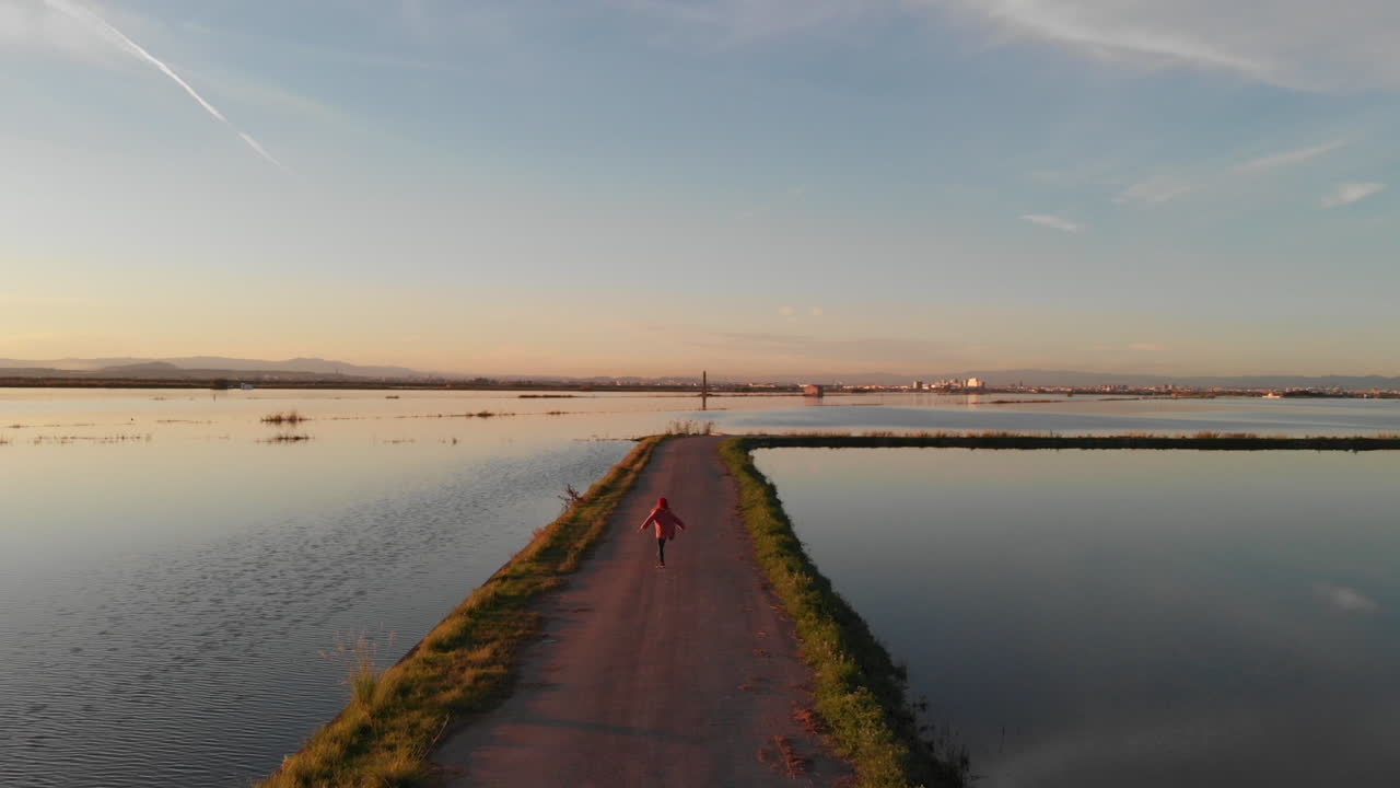 Aerial view following. Beautiful sunset over the rice field covered with water. Perfectly smooth surface of water in Albufera National park, Valencia Spain. Happy Young girl running and jumping
