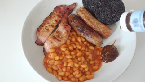 brown sauce being added to a traditional english breakfast of cumberland sausages, back bacon black pudding and baked beans, but no egg, on a white plate