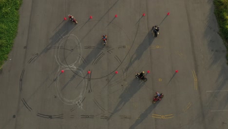 amplia vista aérea de arriba hacia abajo de varios motociclistas practicando en un curso de slalom de entrenamiento avanzado de motocicletas entre conos naranjas con largas sombras y marcas de neumáticos.