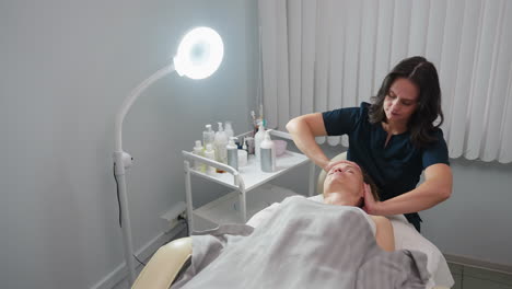 skin therapist tilts head of visitor lying relaxed on treatment bed while massaging neck and scalp with soothing fingers to release tension and promote wellness in serene spa clinic environment