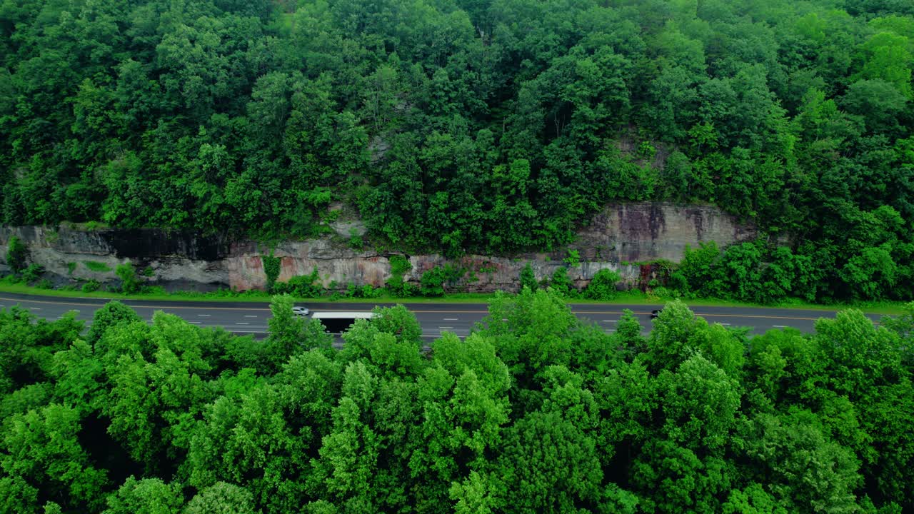 A red Conestoga semi-truck drives along I-24 in Tennessee, cutting through dense forest and rocky cliffside along a rural mountain pass