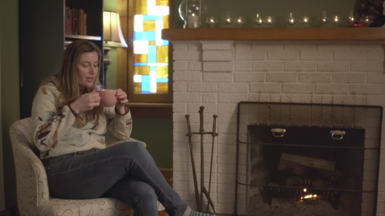 Fireside woman walking in, sitting, relaxing, and drinking tea in a cozy fall setting. She smiles and enjoys the fireplace and atmosphere while drinking a mug of tea. 60 fps Frame Rate, slow motion