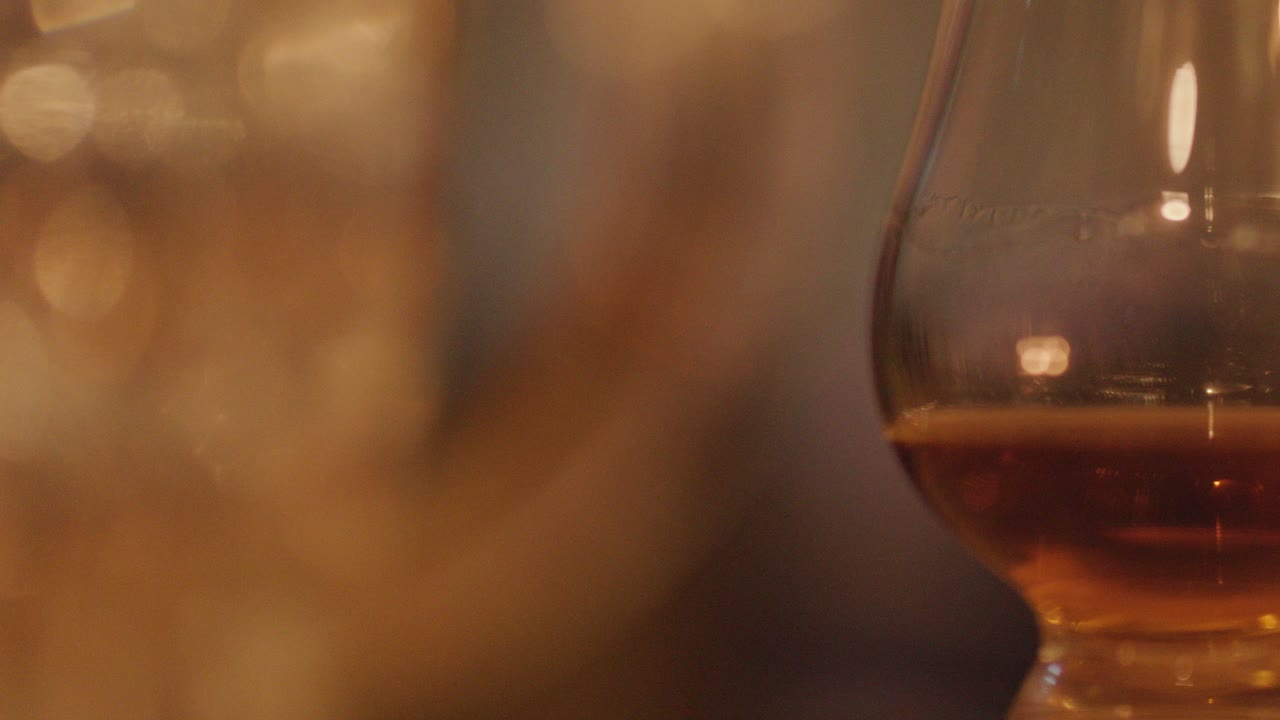 A slow, close-up pan of whisky glasses filled with amber liquid, set against a softly blurred, warmly lit background with candlelight reflections