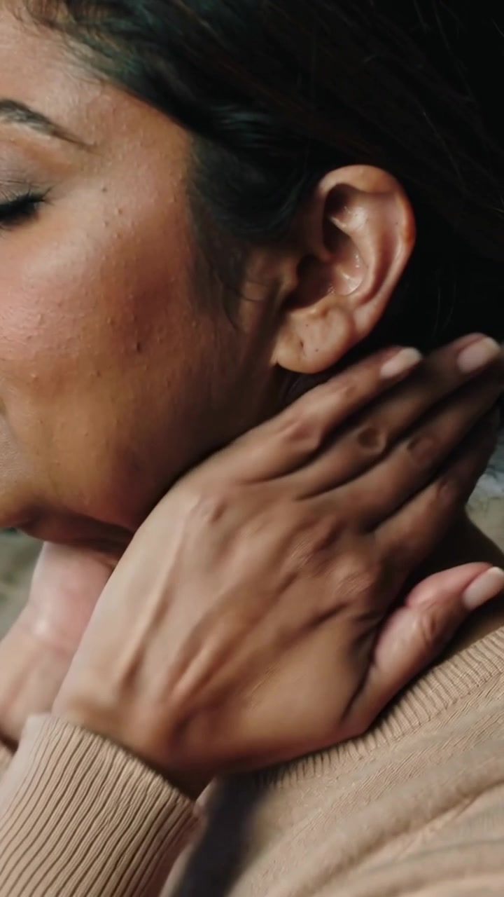 Video verticale: Una donna con un maglione di maglia beige si massaggia il collo a casa, sentendo tensione, spazio per il testo.