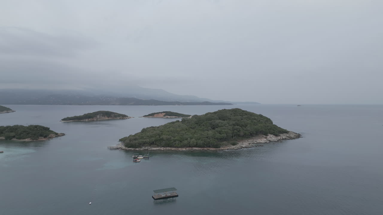 드론이 물 위를 날아서 크사밀 알바니아 앞의 작은 섬으로 날아가는 사진입니다. 구름이 많은 회색 날 아침에 배가 주변에 있고 하늘에는 안개가 있습니다.