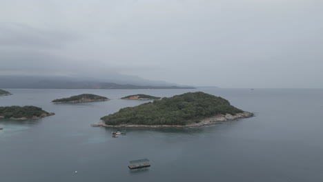 drone shot flying over the water to the small islands in front of ksamil albania in the morning on a cloudy grey day with boats around and haze in the sky log