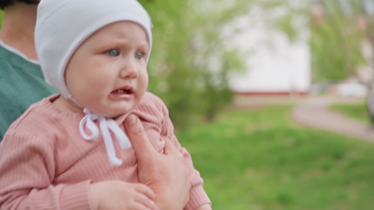 Child Cries Outdoors, Infant Exhibits Distress While Held By Caregiver In Park Setting, Distressed Infant Expresses Discomfort In Caregiver Arms Amidst Park Greenery With Family Close By