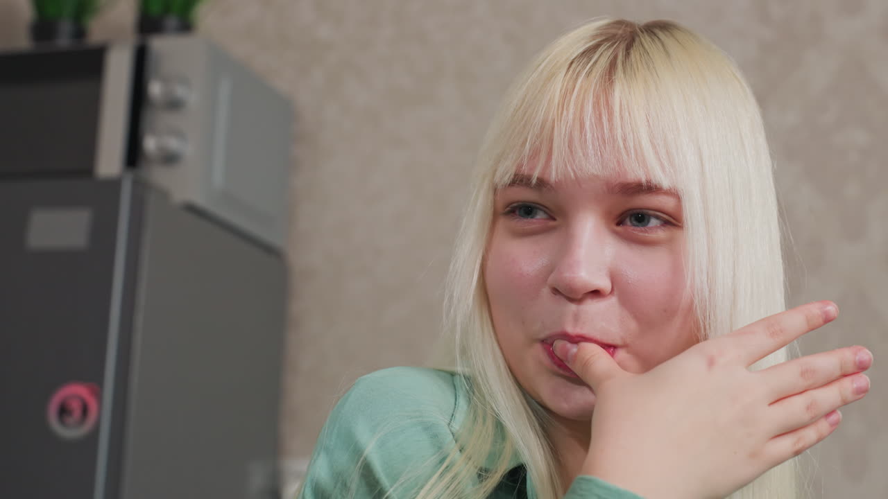 close up of happy woman with blond hair licking finger after eating something, indoor kitchen setting with fridge and microwave in background, joyful moment enjoying snack
