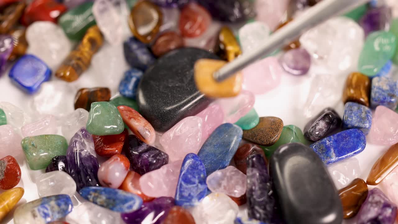 Tweezers pick a tiger's eye stone from a vibrant collection of chakra stones under bright lighting