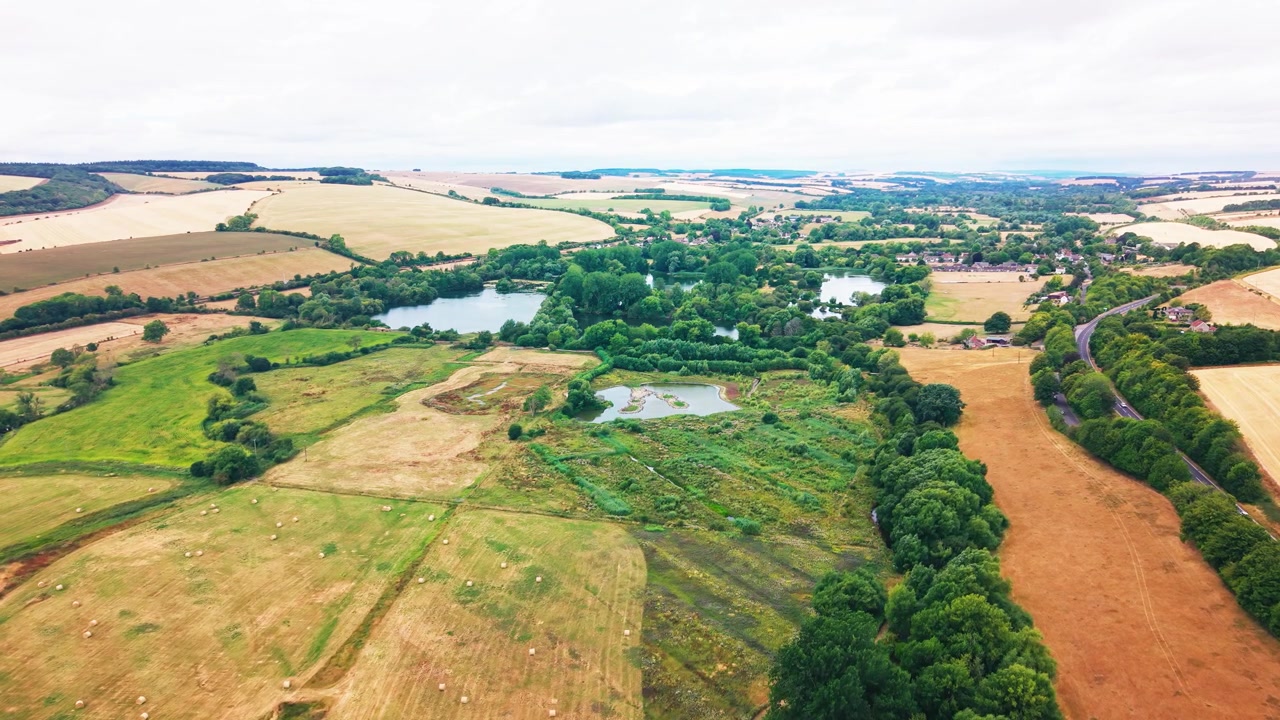 Ampio filmato drone di un villaggio inglese immerso tra laghi e boschi, circondato da un mosaico di campi coltivati e arati con una strada di campagna tortuosa.