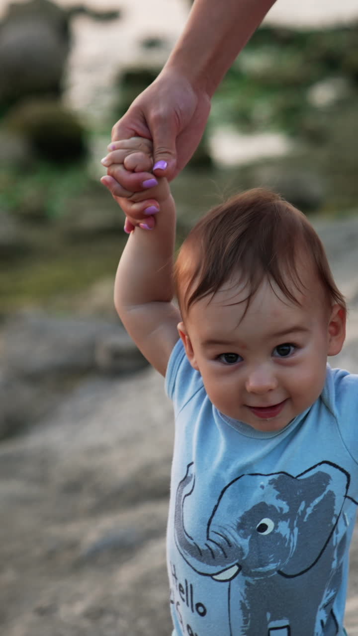 Beautiful Caucasian child walking smiling happily. Mother's hands supporting her baby boy. Nature backdrop in blur. Vertical video