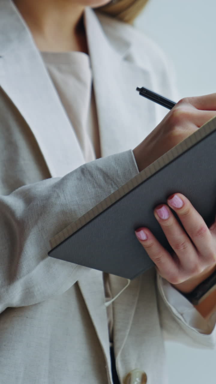 Lady in light jacket standing at the window and holding paper notebook. Woman making notes in her diary. Close up. Vertical video