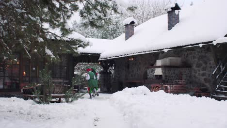 girl and boy elves play with white snow. elves run away to santa claus's wooden house