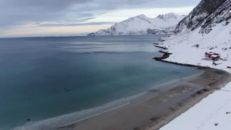 Drone-view-in-Tromso-area-in-winter-flying-over-a-snowy-landscape,-a-brown-beach-and-the-ocean-full-of-snow-in-Norway