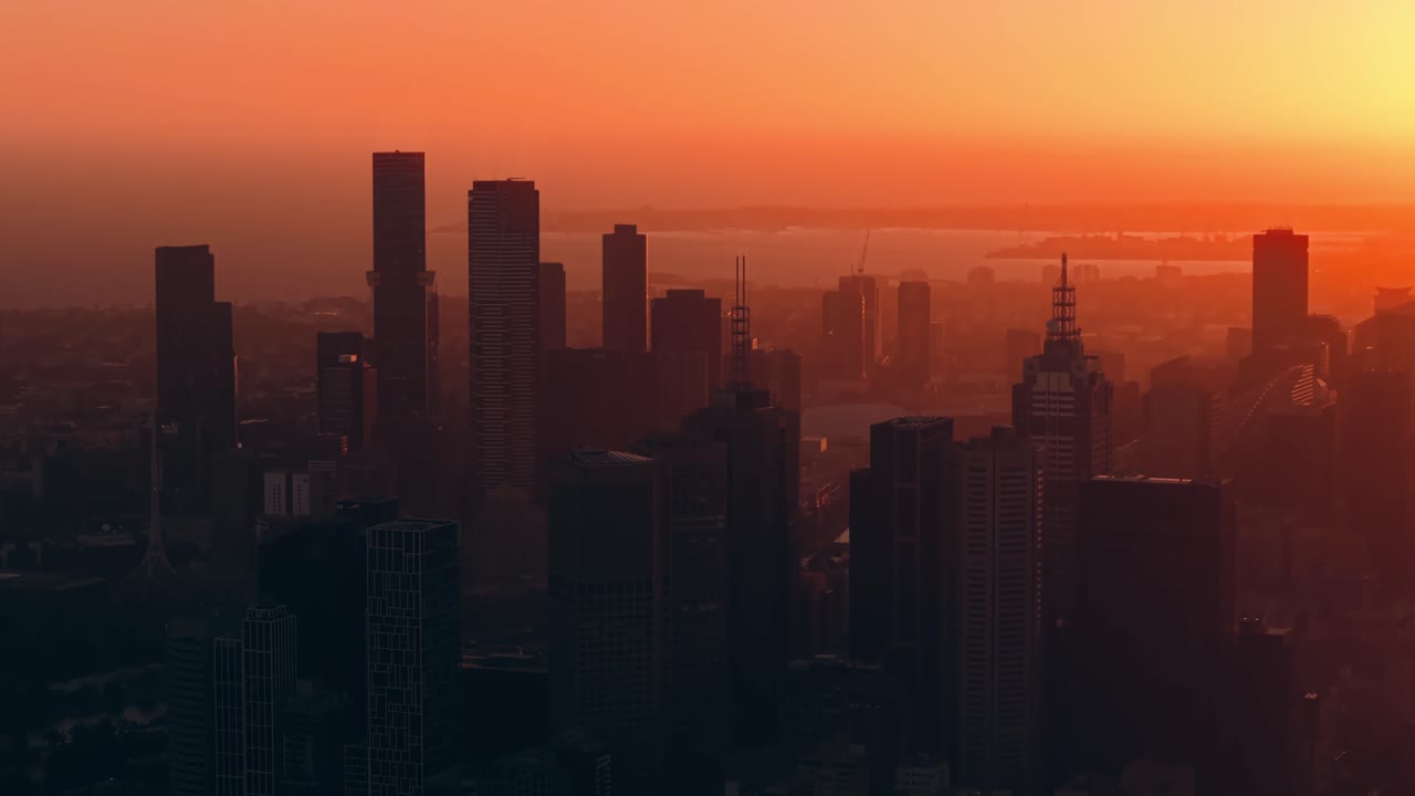Atardecer naranja intenso sobre el CBD de Melbourne con siluetas oscurecidas de rascacielos y luz diurna desvaneciéndose en el horizonte, vista aérea media en ascenso