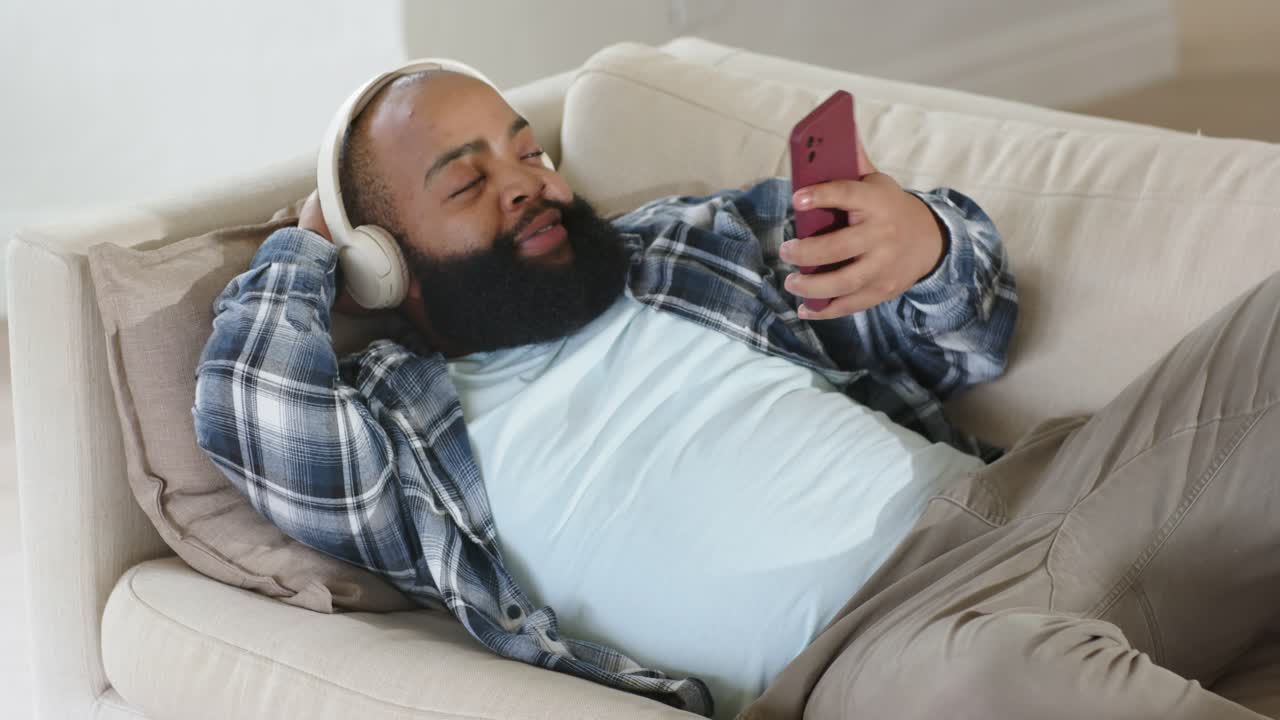 Uomo africano adulto con le cuffie che tiene un telefono rosso e scorre, poi si riposa, rilassandosi sul divano.