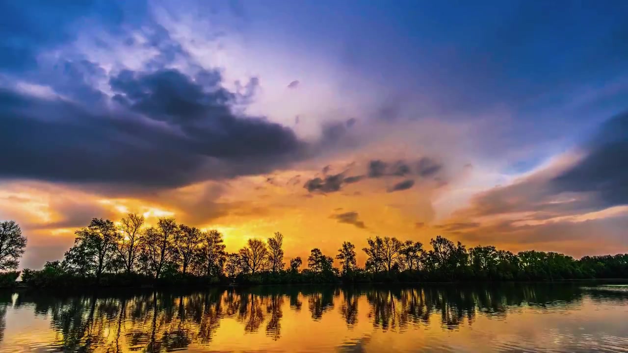 A dramatic sunset time-lapse capturing turbulent stormy clouds reflecting in the calm, mirror-like waters of a tranquil lake with tree silhouettes.