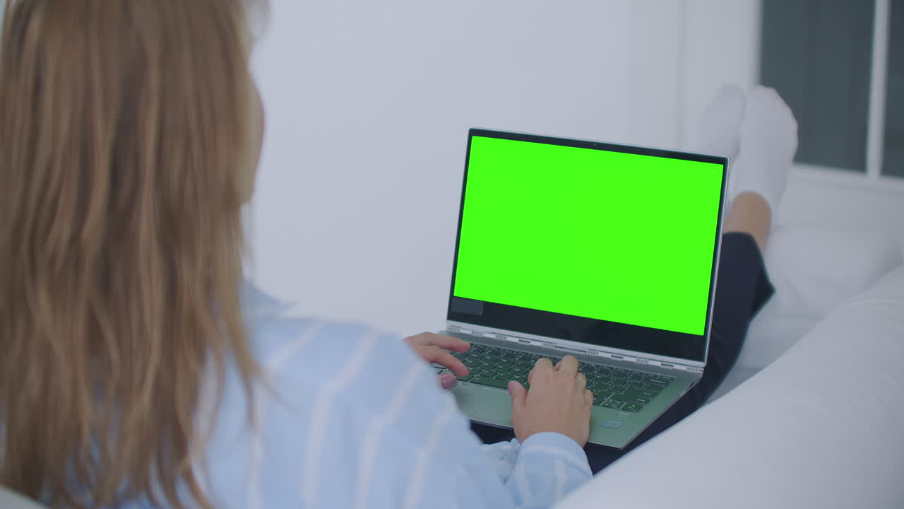 una mujer joven acostada en el sofá mira la pantalla de la computadora portátil con una pantalla verde y asiente con la cabeza. haga una videollamada