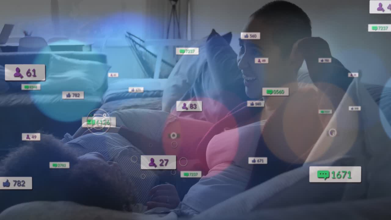 Animation of social media icons over diverse lesbian couple mother embracing each other on the couch