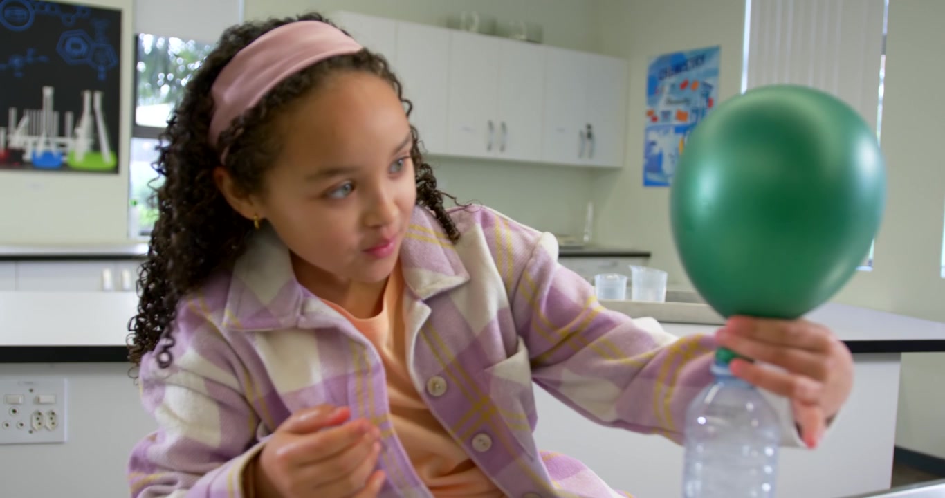 African American girl with pink band steadying bottle as green balloon inflating in lab, to see