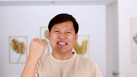 asian man in beige shirt smiles and raises fist in encouragement, standing in a bright, modern room with framed art and soft lighting