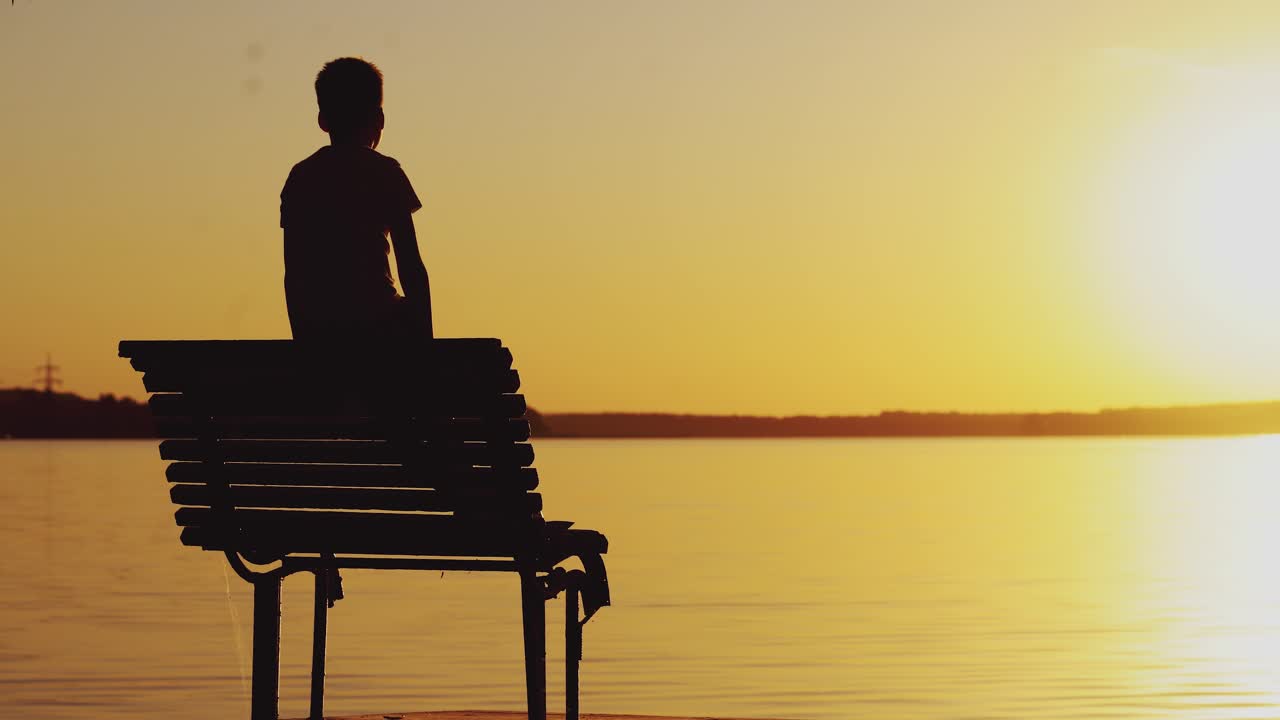 A little boy is looking at the sunset and wonderful landscape near the river on the bench. A guy raises his hands up. Weekend.