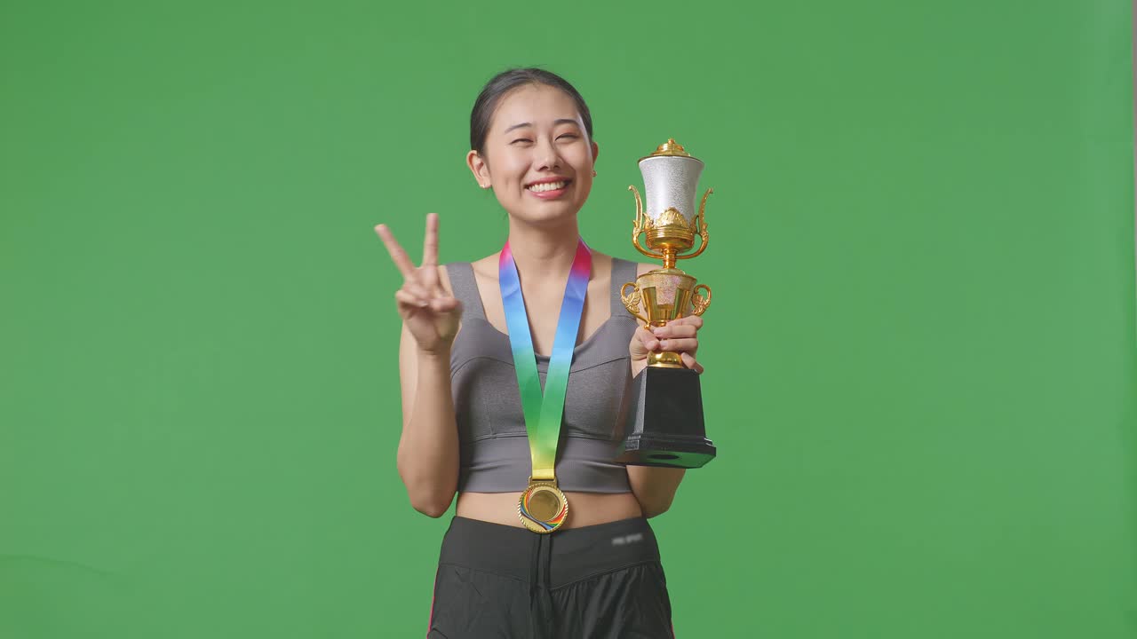 금메달과 트로피를 가진 아시아 여성은 평화의 몸짓을 보여주고 스튜디오의 녹색 스크린 배경에서 첫 번째 우승자로 카메라에 미소 짓고 있습니다.