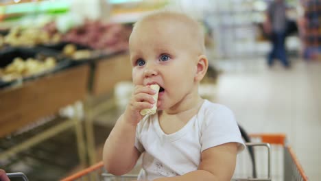 Cute-little-baby-is-taking-banana-and-sitting-in-a-shopping-cart,-while-her-mother-is-pushing-the-cart-forward-walking-in-fruits