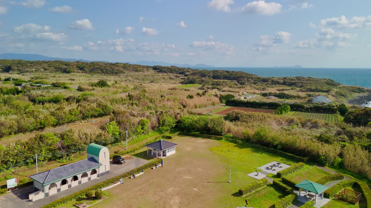 A beautiful aerial push-in shot of the historic Tsunoshima Lighthouse on a scenic coastal hill in Japan, overlooking the ocean under a partly cloudy sky