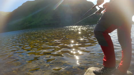 toma estática baja de una persona que lanza una línea de pesca a un río y luego la enrolla, con una montaña en el fondo y una llamarada solar con luz que brilla en el agua