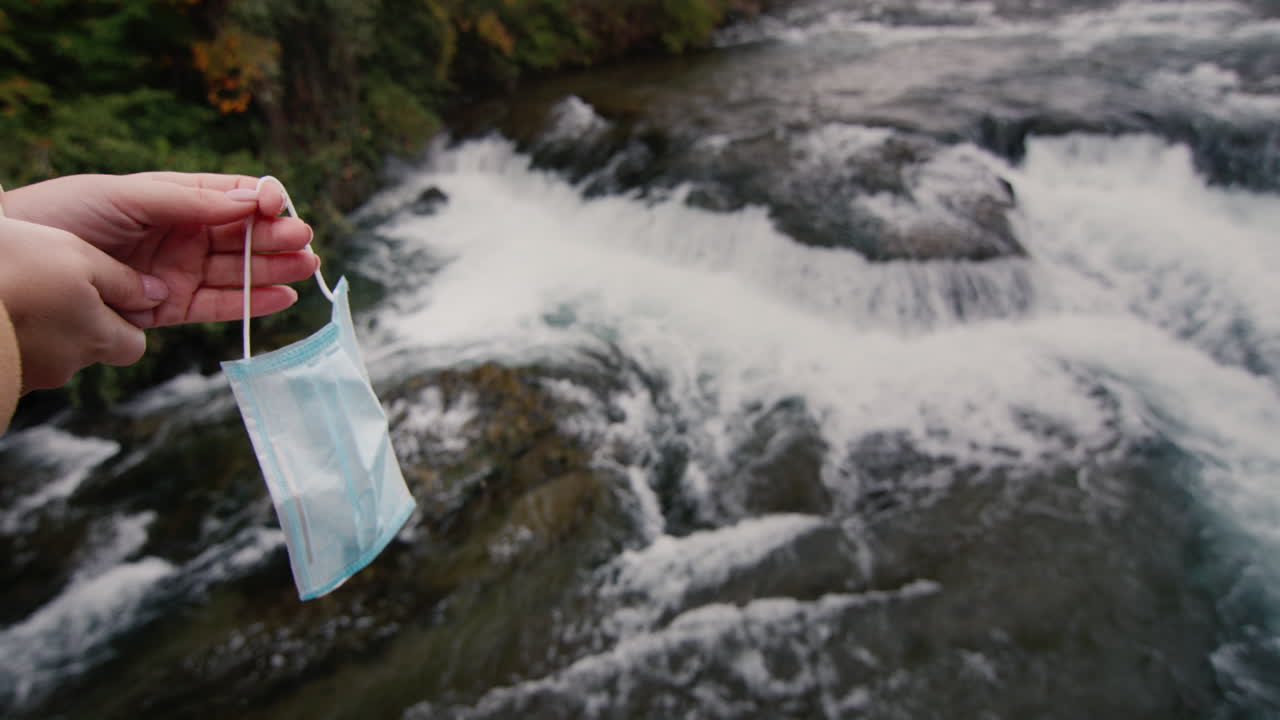 A woman's hand holds a medical mask over a raging mountain river