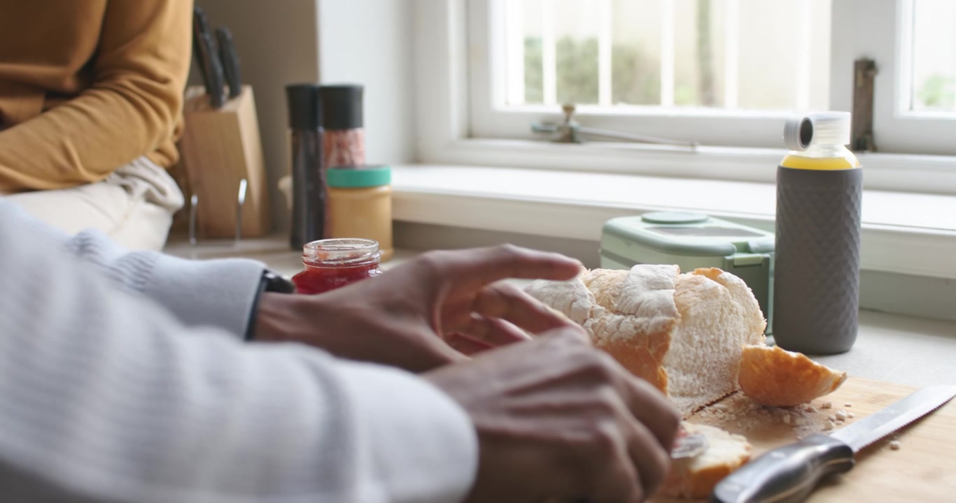 Hånd løsner syltetøylokk og sprer syltetøy på skivet brød ved vindusbenken til frokost.