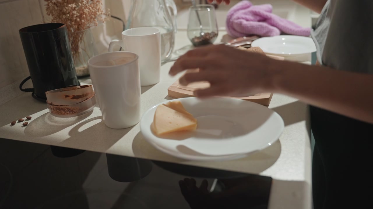 Bancone della cucina illuminato dal sole, donna caucasica che prepara la colazione, amante della musica che si gode un rituale calmo, mani che affettano pane integrale, formaggio su piatto bianco, tazza di ceramica, bagliore caldo, fiori secchi, casa accogliente.
