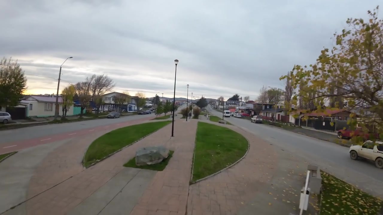 Spartitraffico alberato e rotatoria su Avenida Santiago Bueras, con case colorate e basse colline sotto un cielo nuvoloso a Puerto Natales, Cile, ripresa aerea FPV