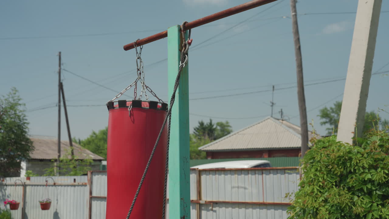 Red Punching Bag Hanging From Wooden Pole In Backyard, Chained Support And Rustic Fence Under Clear Sky, Outdoor Training Prop With Textured Surface, Quiet Summer Scene With Suburban Houses