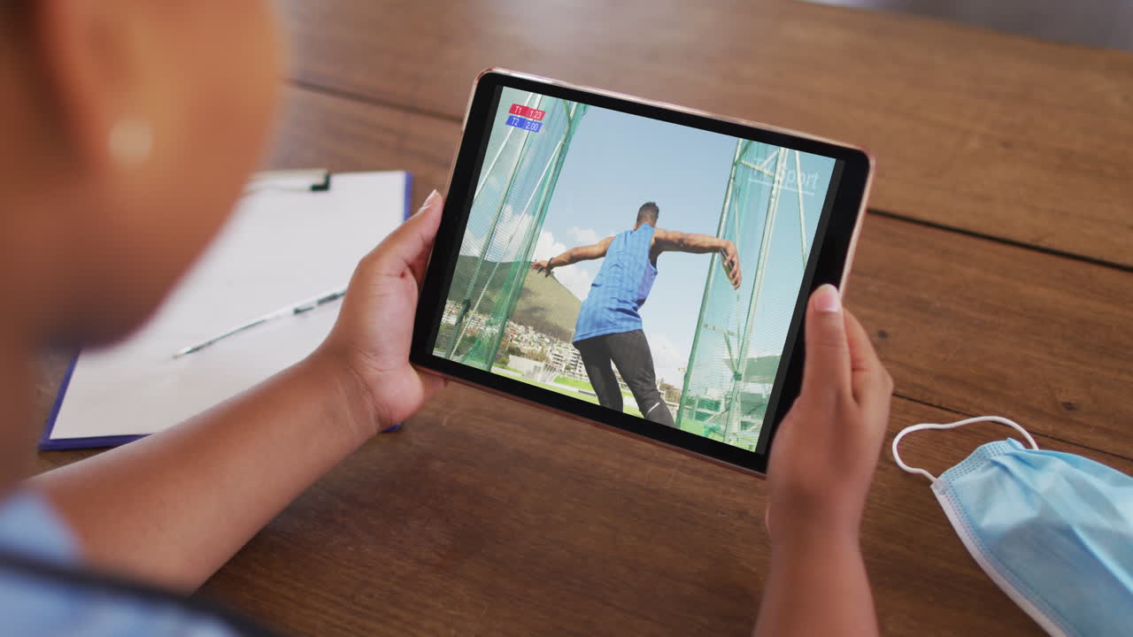 태블릿에서 육상 디스크 이벤트를 시청하는 얼굴 마스크와 함께 테이블에 앉아있는 여성의 합성