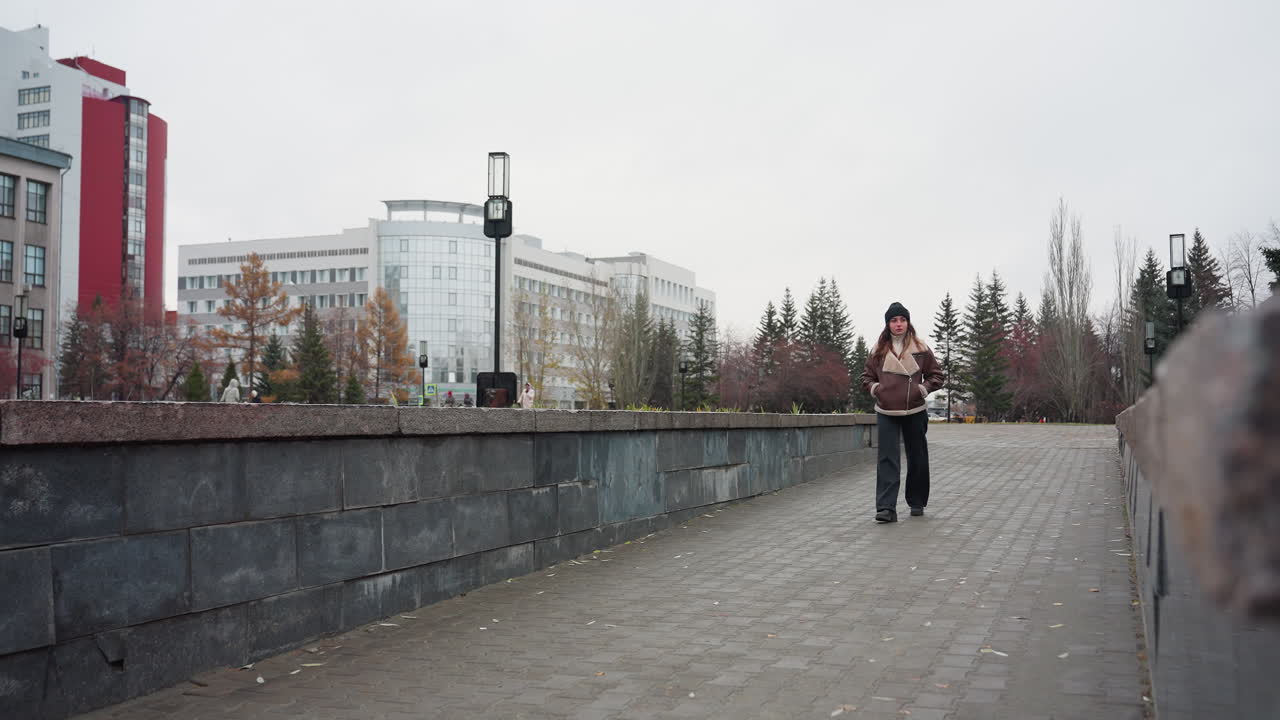 Student in black cap brown jacket walking slowly hands in pockets on wide stone pathway through urban park surrounded by pine trees modern buildings and cloudy sky in calm peaceful autumn atmosphere