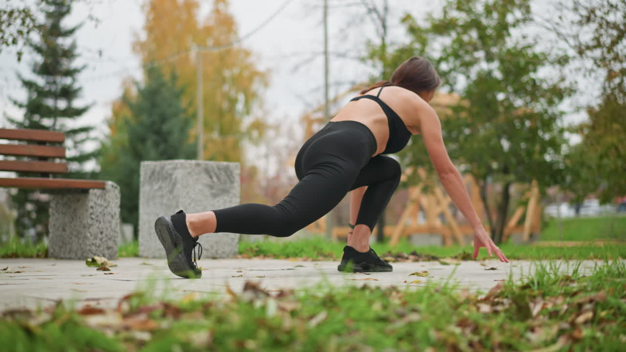 A woman stretches out her legs, getting ready to run, with her left leg forward and right leg back, the background features a concrete bench and trees, setting an outdoor athletic vibe