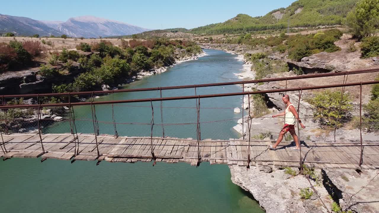 un turista camina a través de un puente colgante de madera y tambaleante en el río vjosa, albania - camiones aéreos de lado