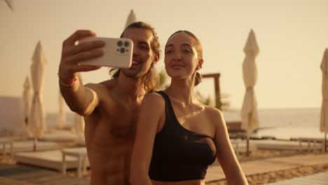 a guy and a girl in a black top take a selfie against the backdrop of a sunny golden beach with folded umbrellas and palm trees