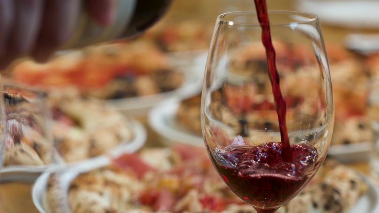 Primo piano di vino rosso che si versa in un bicchiere con pizze sullo sfondo sfocato
