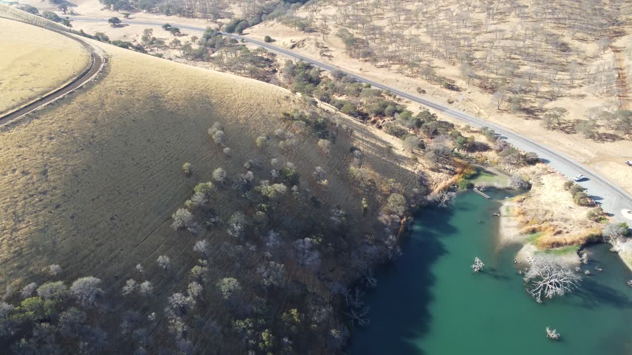 High flying drone footage of small water mass tucked away in between golden hills, road traveling through