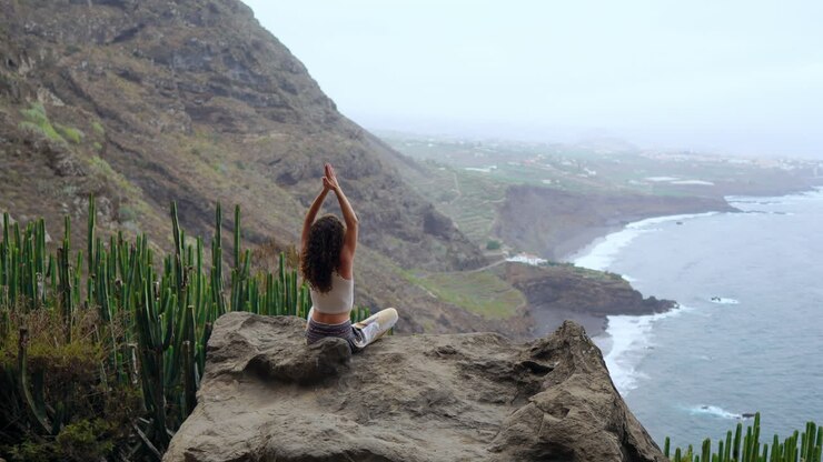 Practicing yoga amidst island mountains, a young woman sits on a mountaintop rock, meditating in Lotus posture, with a view of the ocean below