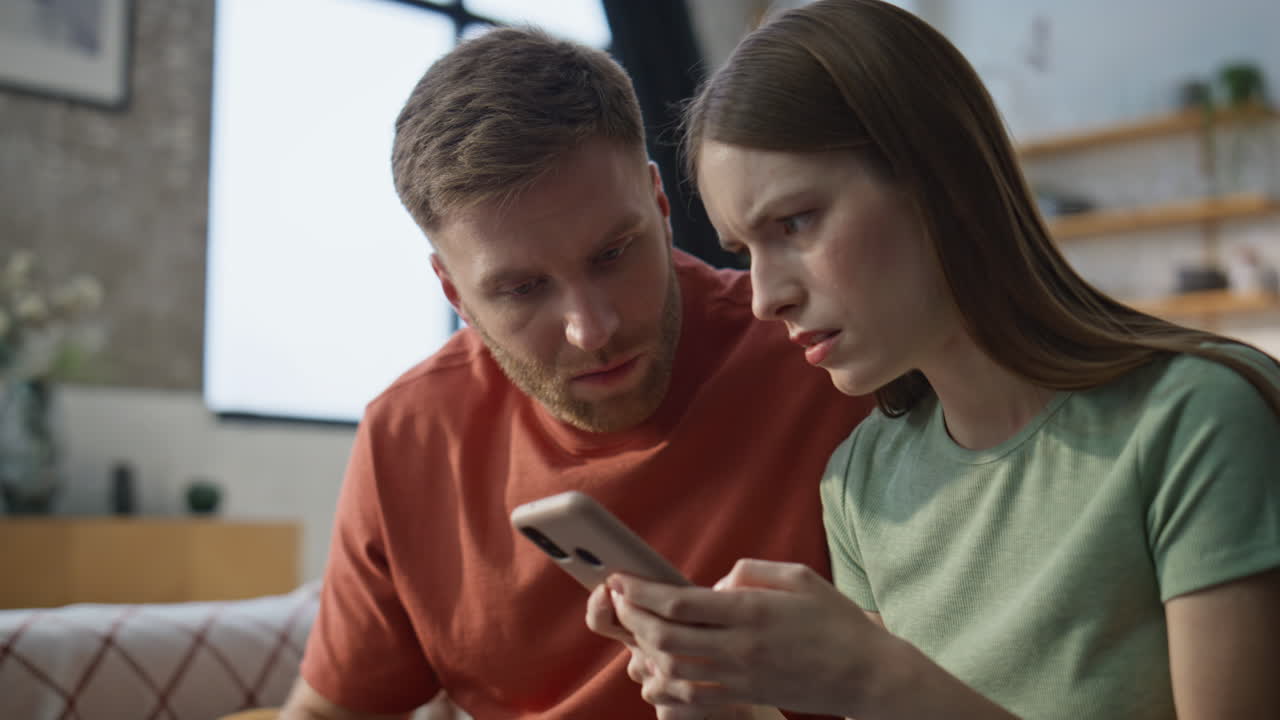 Worried wife showing message smartphone to husband sitting at couch closeup